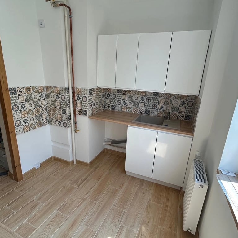 Mała kuchnia po remoncie z białymi szafkami, drewnianym blatem i dekoracyjnymi płytkami w stylu patchwork, jasna podłoga imit