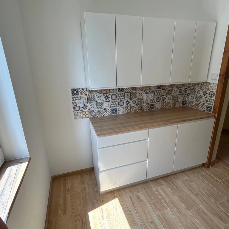 Kuchnia po remoncie z białymi szafkami, drewnianym blatem i pralką pod zabudową, ściana wykończona kolorowymi płytkami patchw