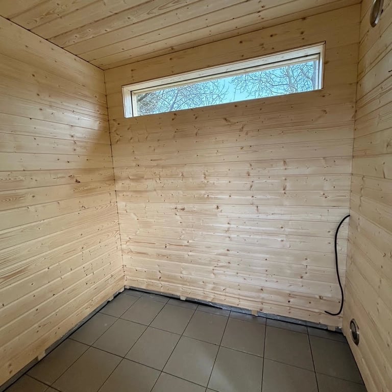 Rozbudowa domu z częścią sauny, elewacja drewniana z poziomymi deskami, okno z czarną ramą, ogród w tle.