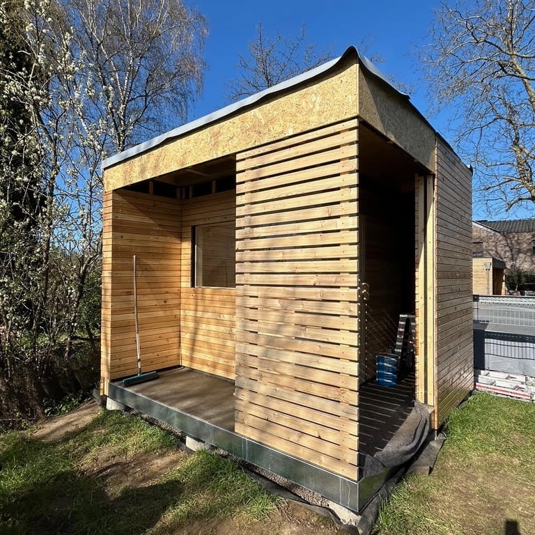 Sauna ogrodowa w trakcie budowy, drewniana elewacja z pionowymi deskami, drzwi z przeszkleniem i otwór okienny.