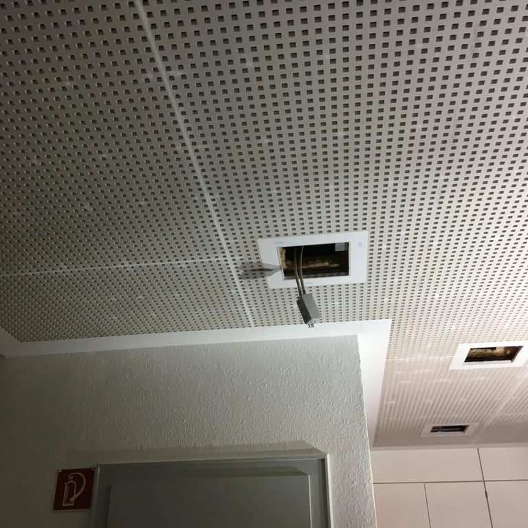 Sufit akustyczny z perforowanymi płytami, otwory na lampy i instalacje elektryczne, jasne ściany w tle