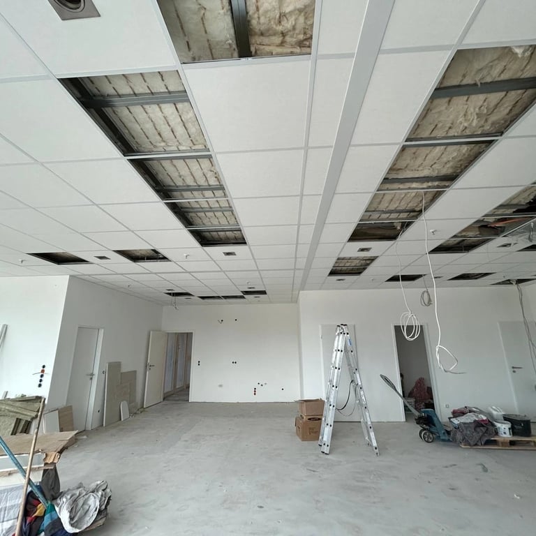 Montaż sufitu kasetonowego w biurze, widoczne drabiny, materiały budowlane i konstrukcja stelaża pod kasetony.