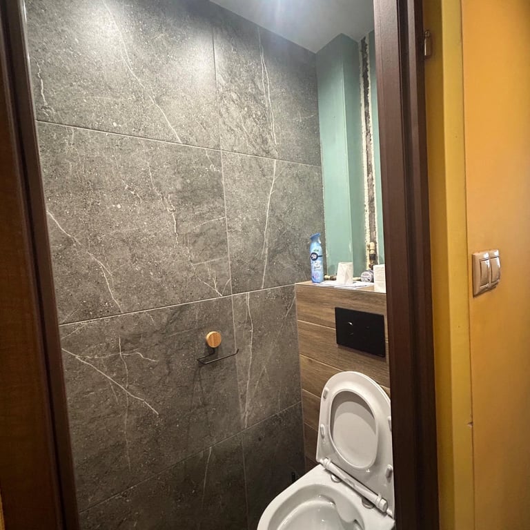 Toaleta z beżowymi płytkami na ścianach, zabudowa stelaża podtynkowego, oświetlenie LED w suficie i chromowana spłuczka.