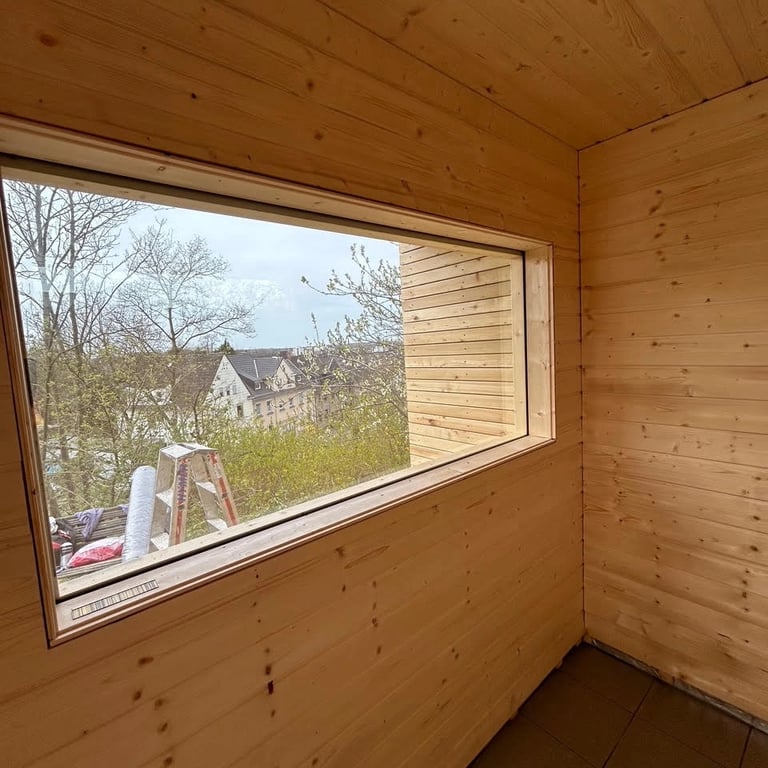 Wnętrze sauny z dużym panoramicznym oknem, drewniane ściany i sufit, podłoga z płytek ceramicznych.