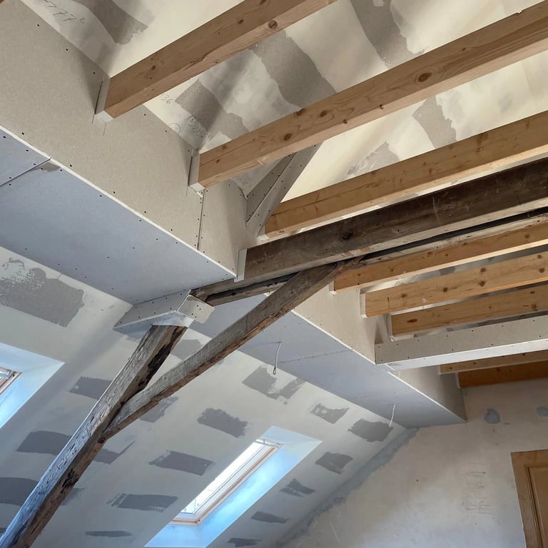 Wykończenie poddasza z płytami gipsowo-kartonowymi, widoczne drewniane belki więźby dachowej i okna dachowe.