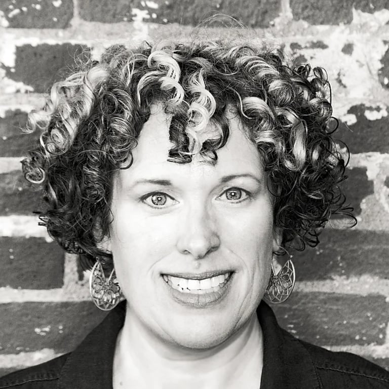 Heather Blaikie, a woman with curly hair and a black jacket standing in front of a brick wall