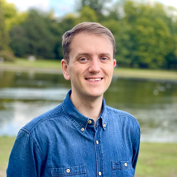 Chris Philpot, a white man in his 30s, smiling at the camera. He is wearing a blue denim shirt and standing in a park.