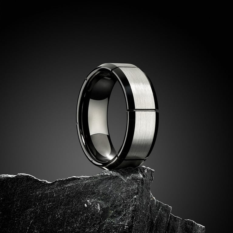 A professional product photo of a two-tone ring with a brushed silver center and polished black edges, resting on a dark rock