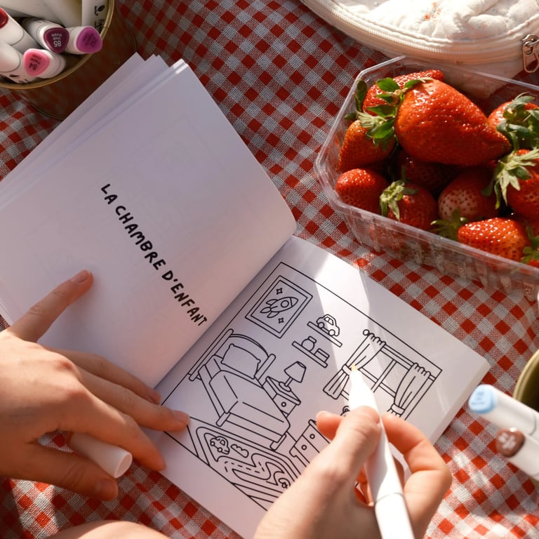 Une main coloriant une scène de chambre dans un livre de coloriage, à côté de fraises fraîches