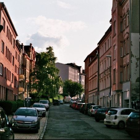 Narrow European city street lined with parked cars and classic brick residential apartment buildings.