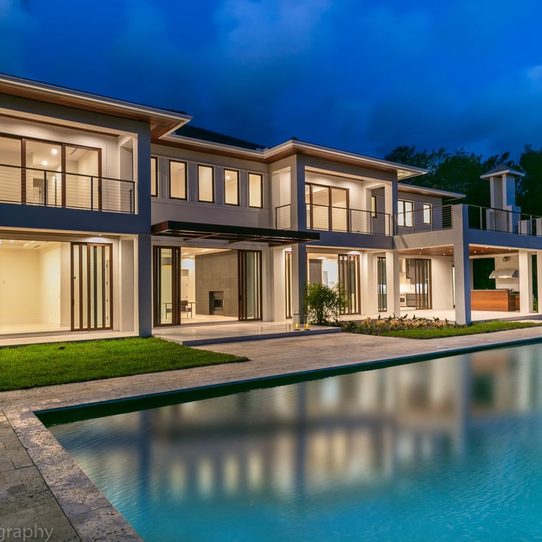 the backyard of a large luxury home showing the back patio and pool at night lit up