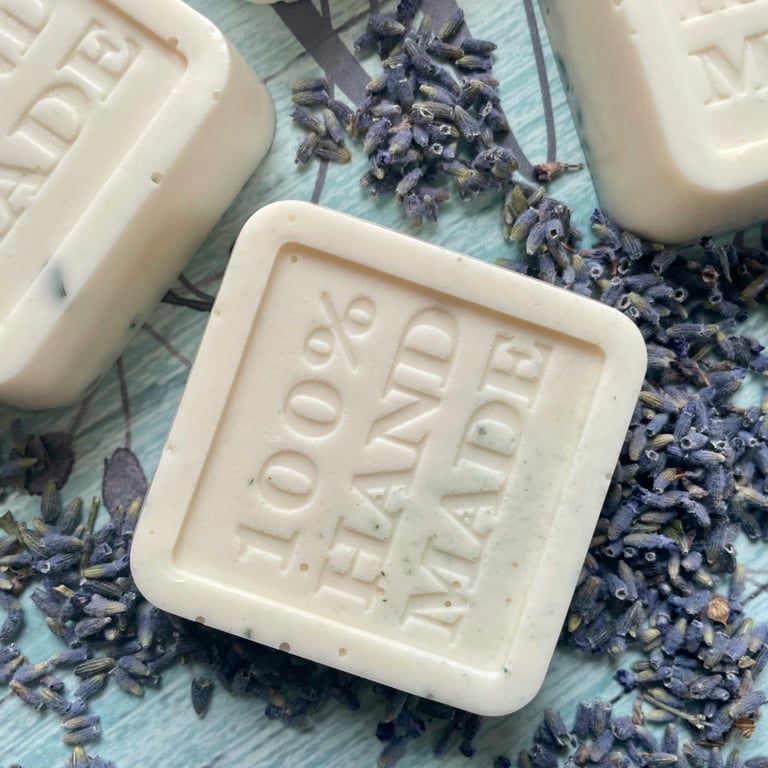 Square white 100% handmade soap bars with embossed text and dried lavender buds on a blue surface.