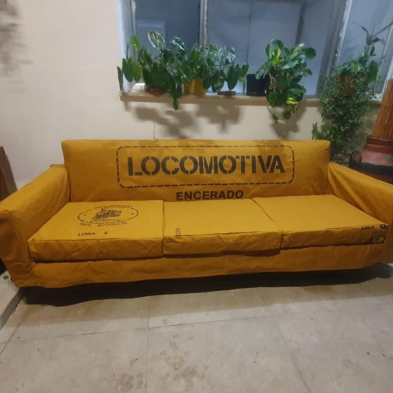 Sofá industrial de lona amarela com a marca Locomotiva Encerado e plantas em vasos ao fundo.