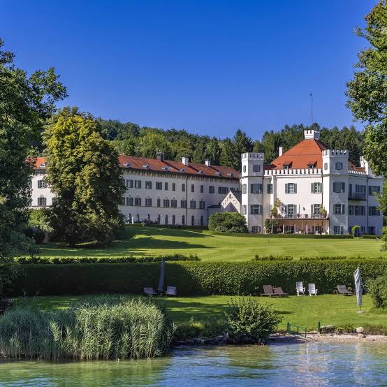 Historic white castle with orange roofs overlooking a green lawn and lakefront at Lake Starnberg.