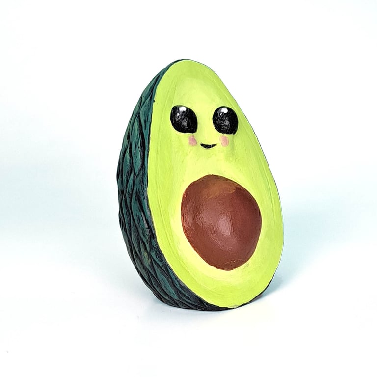 Sculpture of a cute avacado. It has big eyes and rosy lil cheeks and a cute lil smile