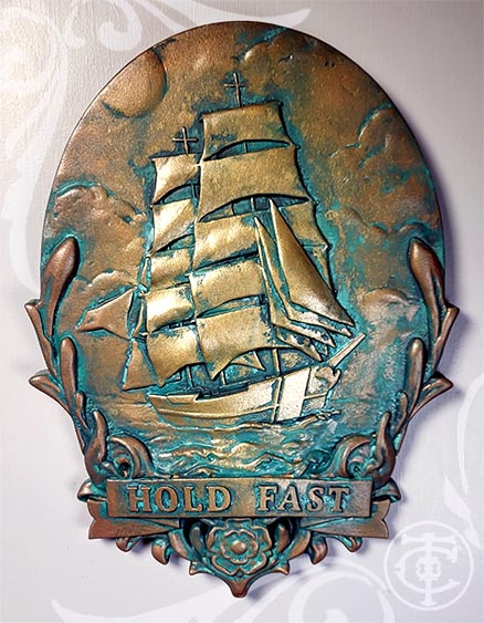 Image of a custom plaque. It shows a tall ship sailing. Below it is a scrollwork frame with a banner  that says Hold Fast. 