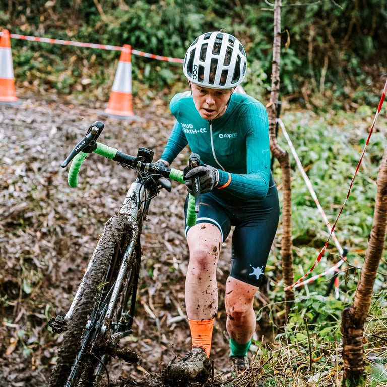 Liz Hughes during a cyclocross race, dismounted and running with her bike through a muddy ditch.