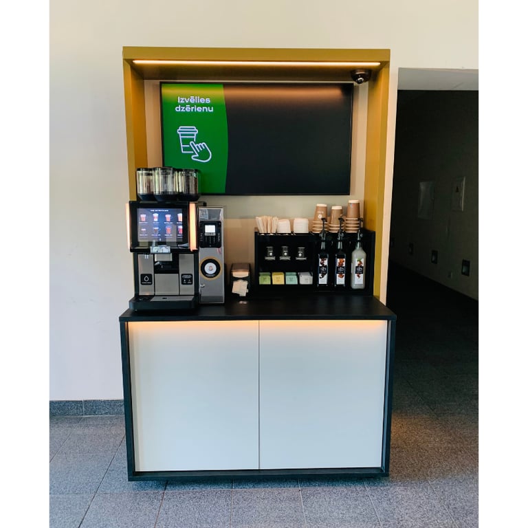 Pašapkalpošanās kafijas stacija McReil birojā ar iebūvētu kafijas automātu un uzkodu plauktiem.