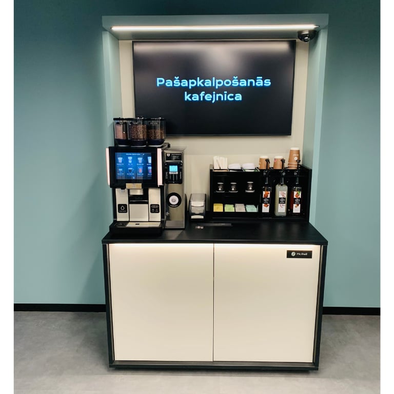 Pašapkalpošanās kafijas stacija McReil birojā ar iebūvētu kafijas automātu un uzkodu plauktiem.