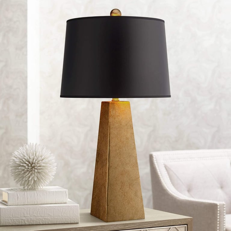 Modern gold pyramid table lamp with a black drum shade on a nightstand for bedroom lighting.