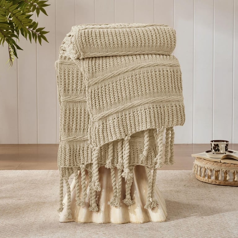 Cream chunky knit throw blanket with tassels draped over a stool in a cozy living room.
