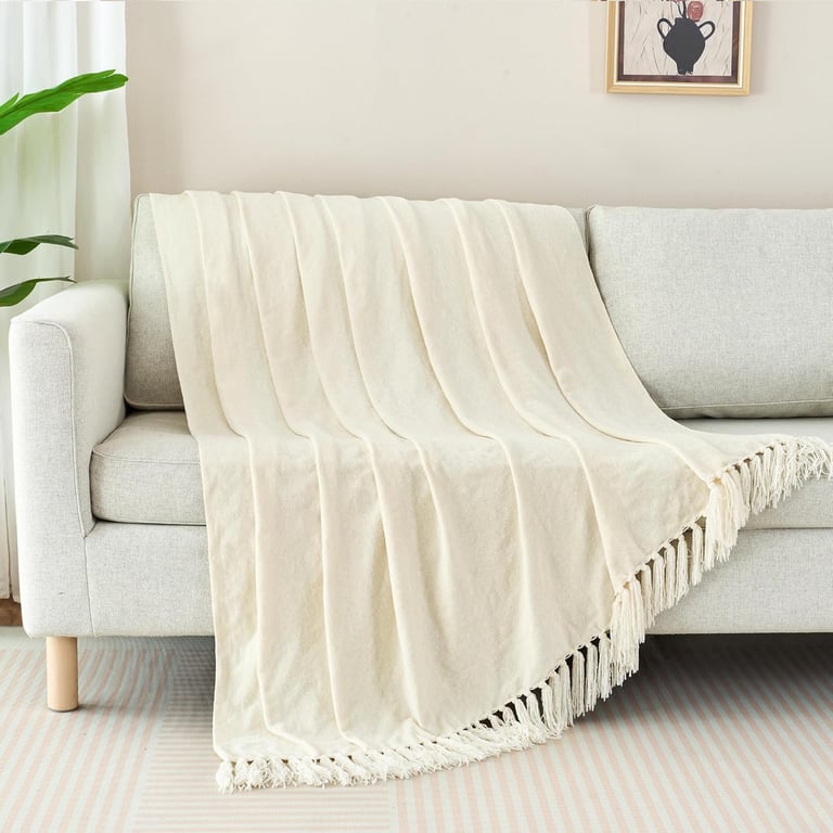 Cream-colored woven throw blanket with fringe draped over a modern light gray sofa in a living room.