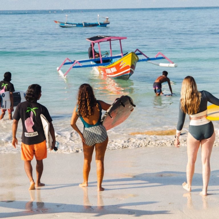 Surfers walking on a white sand beach in Lombok with a colorful outrigger boat in the ocean.