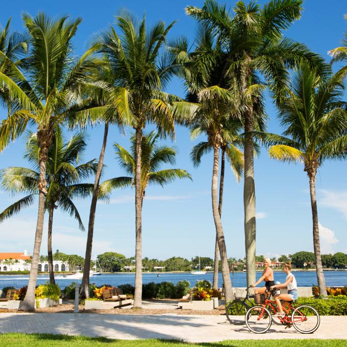 Two women cycling on a tropical waterfront path lined with palm trees in West Palm Beach, Florida.