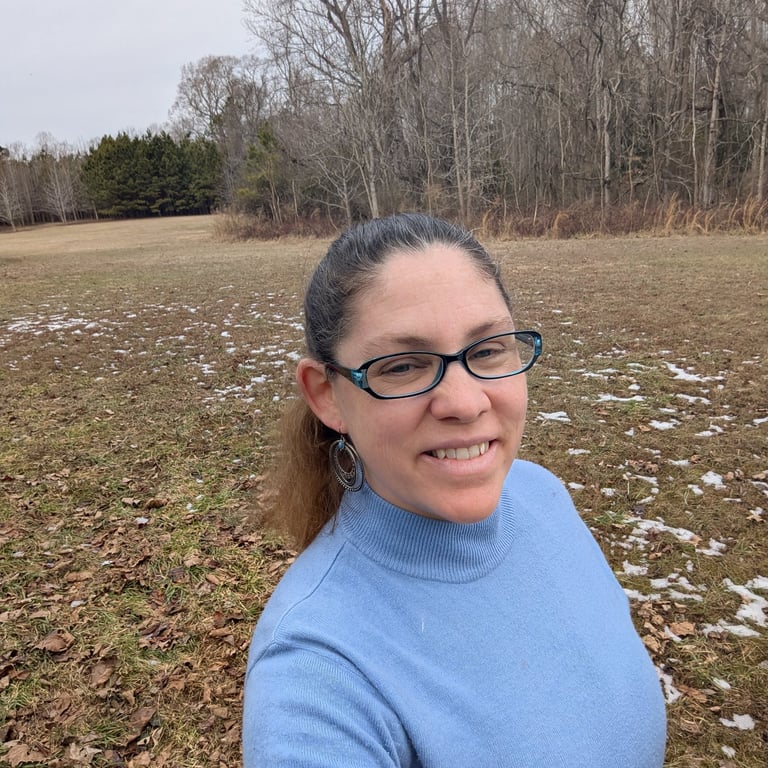 Photo of me, a white woman with long brown hair in a ponytail, wearing a blue shirt & glasses. Field and trees in background