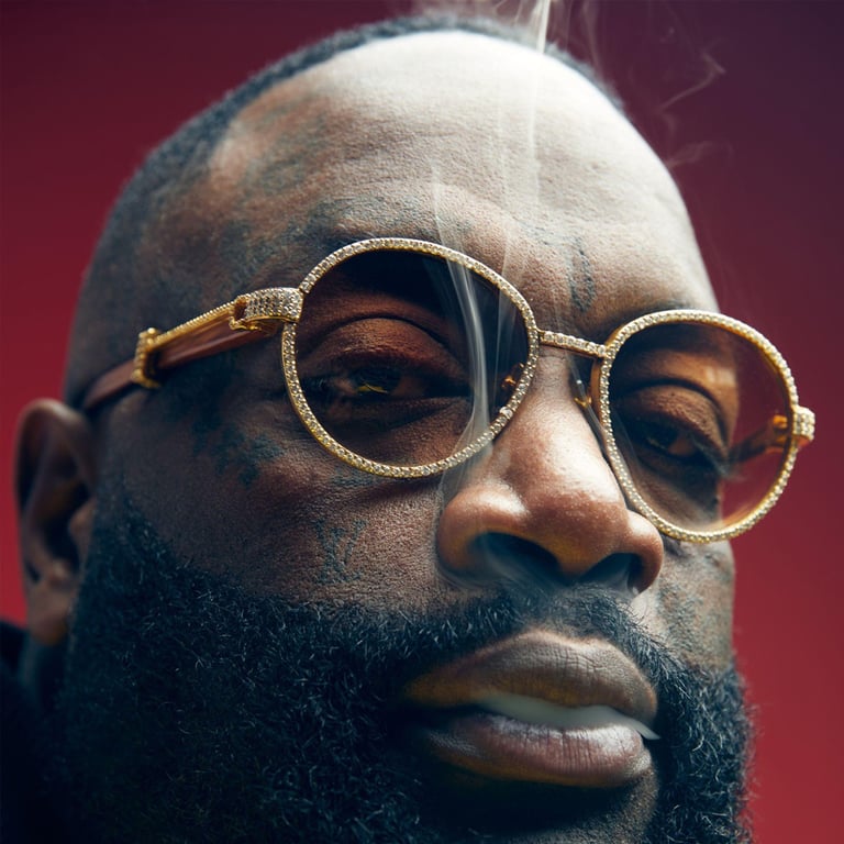 Close-up of Rick Ross wearing diamond-encrusted sunglasses and exhale cigar smoke.