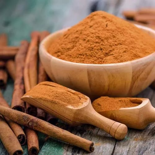 A wooden bowl and scoops filled with ground cinnamon powder next to cinnamon sticks on a rustic table.