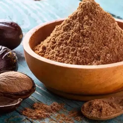A small wooden bowl filled with ground nutmeg powder next to whole nutmeg seeds on a rustic blue table.