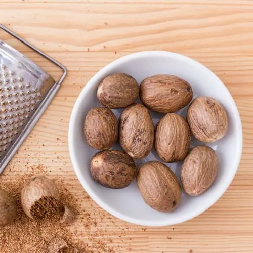 Whole nutmeg seeds in a white bowl next to a metal grater and freshly ground nutmeg on a wooden surface.