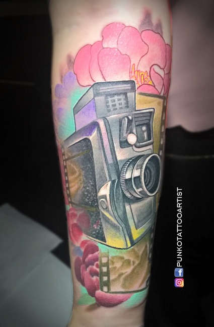 Tatuaje  de **cámara fotográfica antigua** con detalles botánicos en línea fina. Obra realizada en el antebrazo 