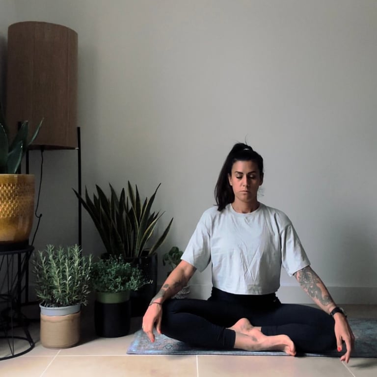 A woman with tattoos practicing mindfulness meditation in a cross-legged yoga pose at home.