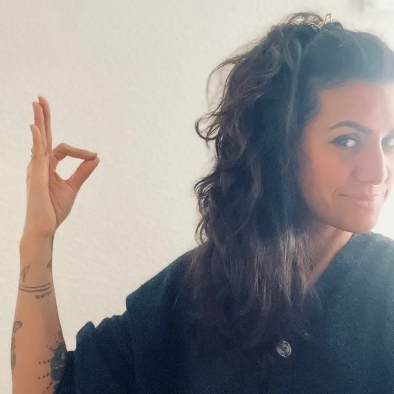 A woman with wavy dark hair and arm tattoos making an okay hand gesture against a plain wall.