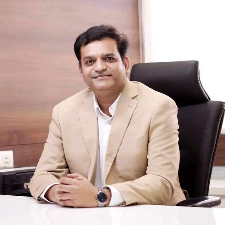 Professional portrait of a male business executive in a beige blazer sitting at an office desk.