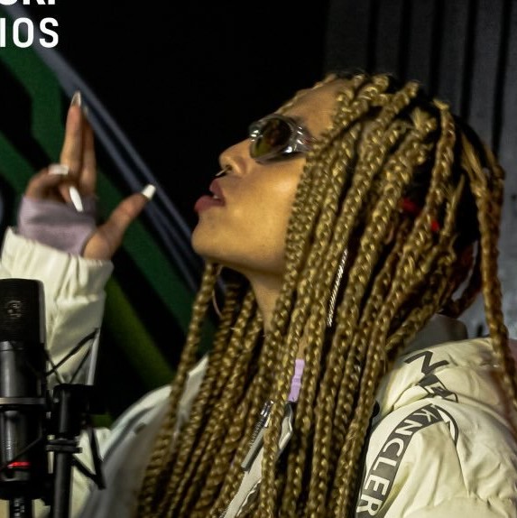 Artista con trenzas boxeadoras rubias, usando gafas de sol y una chaqueta acolchada blanca 