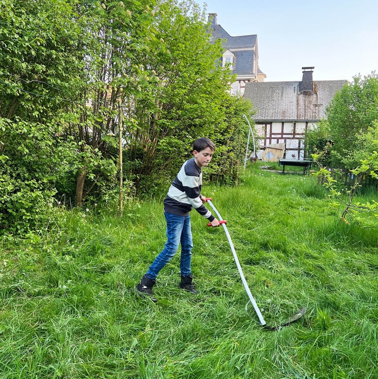 Philipp Geisler mäht mit einer Sense die Wiese in seinem Garten