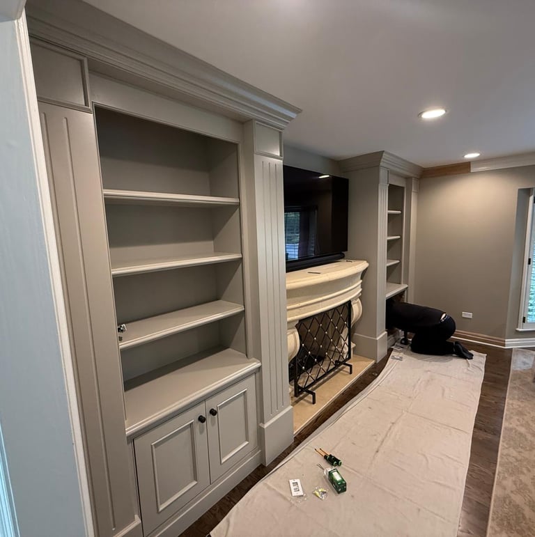 A carpenter installs custom gray built-in bookshelves and cabinetry flanking a living room fireplace.