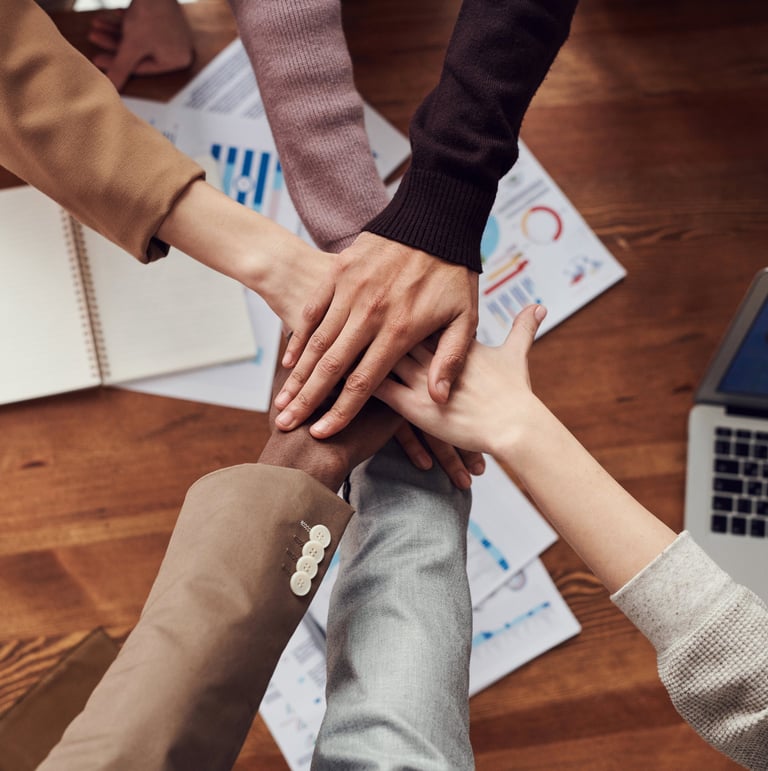 Stacked hands over a desk with charts and a laptop, symbolizing teamwork.