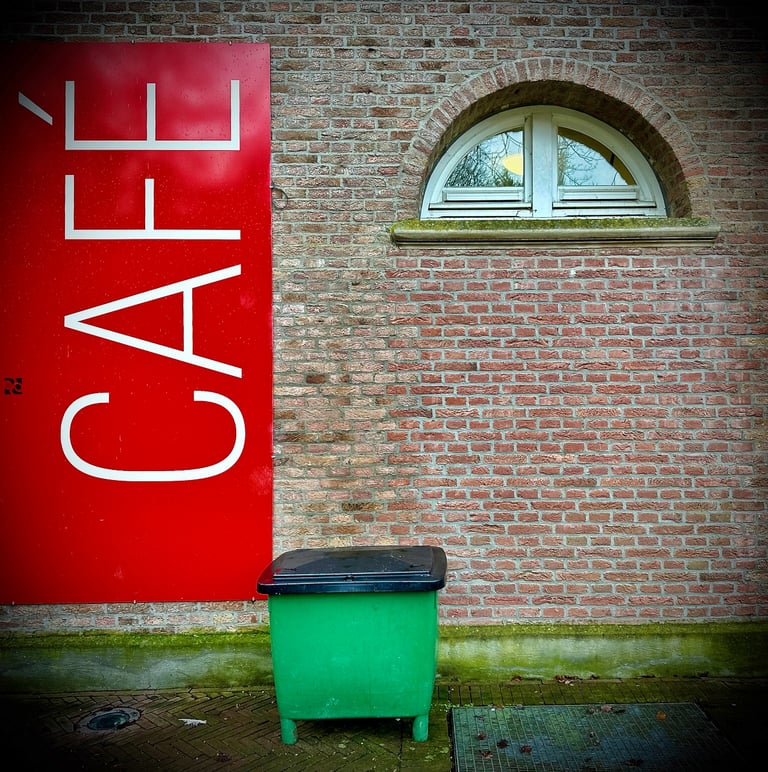 Vertical red cafe sign on a brick wall next to a green trash bin and arched window.