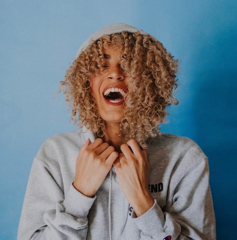Vrouw met krullend haar in een hoodie, lachend en blij.