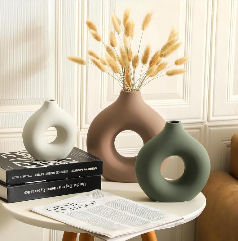 Three modern ceramic donut vases in white, brown, and green with dried pampas grass on a side table.
