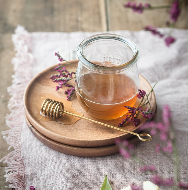 Golden organic honey in a glass jar with a metal dipper and purple flowers on a rustic wooden table.