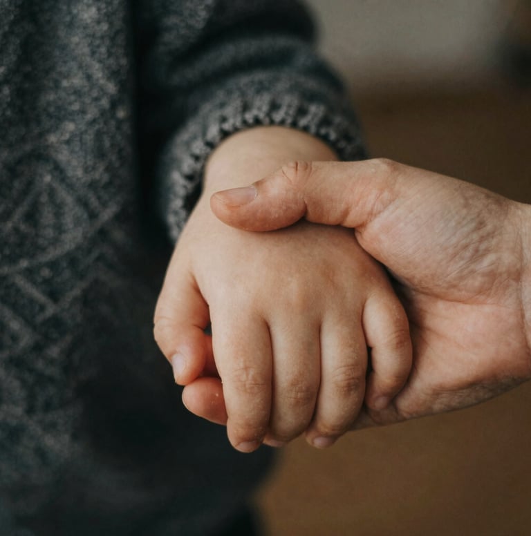 A close-up, candid shot of a young child's hand holding a parent's hand. The lighting is soft and warm, with a shallow depth of field. Soft skin tones against a Charcoal-colored knit sweater. Authentic and intimate.