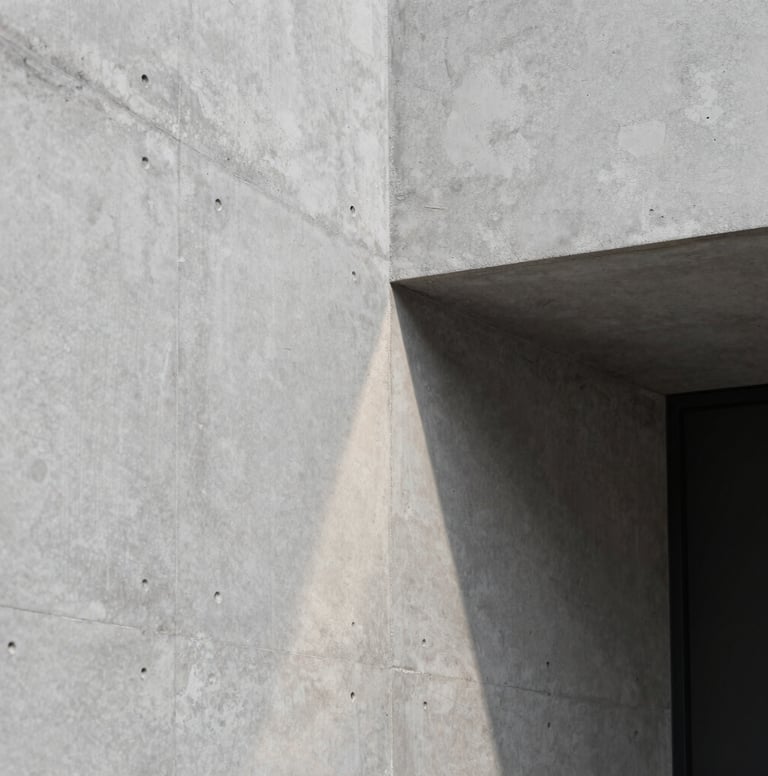 Close-up photography of a polished concrete wall in a Brazilian design studio. A single ray of light hits the surface, revealing smooth texture. Sophisticated and minimalist composition, neutral light grey and black tones.