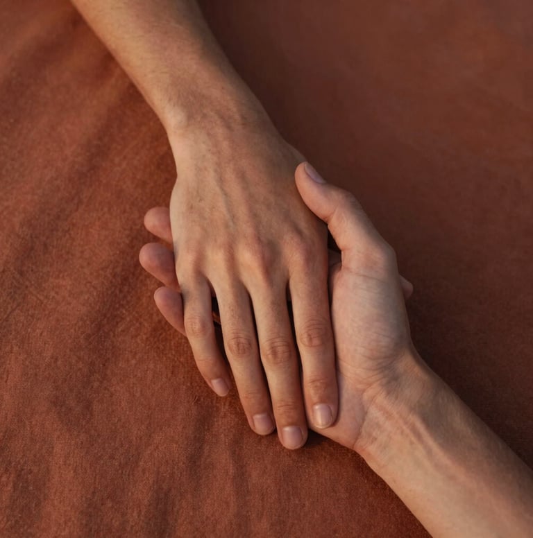 A close-up lifestyle photograph focusing on two hands gently holding each other, with a textured terracotta fabric in the background. Taken in Spain / Southern Europe during the golden hour. The lighting is soft and warm, highlighting the natural skin texture and a sense of deep, intimate connection. Authentic, natural, deeply emotional, cinematic depth of field.