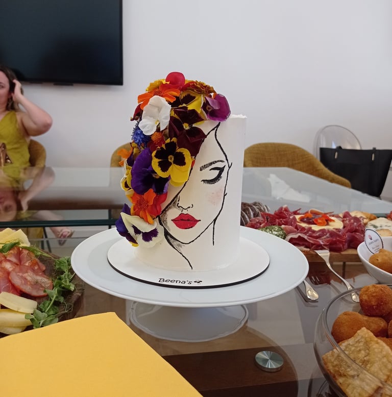 Elegant tall white cake featuring a hand-painted woman's face and colorful edible flowers.