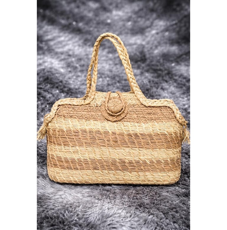 Handwoven straw tote bag with braided handles and horizontal striped texture on a grey faux fur background.
